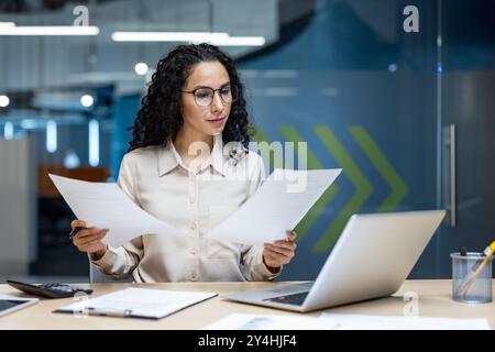 Seriöser hispanischer Buchhalter, der Dokumente am Schreibtisch mit Laptop in heller Büroumgebung überprüft. Frau trägt eine Brille und zeigt sich beruflich auf finanzielle Aufgaben während des Arbeitstages. Stockfoto