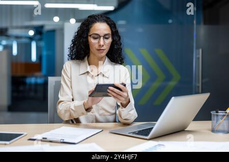 Hispanischer Buchhalter, der mit detaillierten Finanzanalysen beschäftigt ist und den Rechner in der Hand hält. Sie konzentriert sich auf Zahlen und Daten und arbeitet fleißig am Schreibtisch mit Papieren und Laptops. Stockfoto