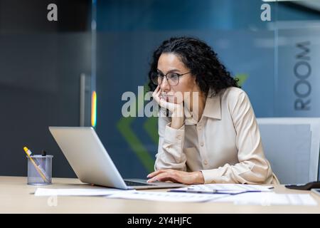 Professionelle Geschäftsfrau mit Brille im Büro mit Laptop. Fokussierter Ausdruck, Analyse von Dokumenten auf dem Schreibtisch. Die Büroumgebung schlägt Rollen wie Manager, Analysten oder Berater vor. Stockfoto