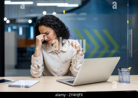 Gestresste Büroangestellte, die die Augen vor dem Laptop reiben, was auf Druck am Arbeitsplatz hinweist. Das Notebook auf dem Schreibtisch weist auf eine geschäftige Umgebung hin. Das Bild vermittelt Spannung, Ermüdung und die Notwendigkeit einer Unterbrechung. Stockfoto