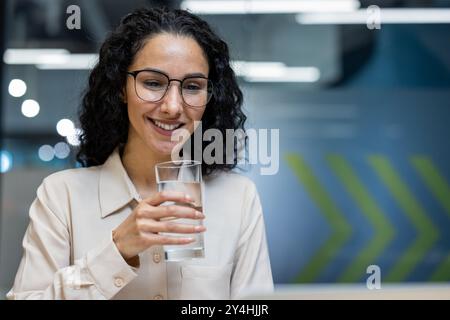 Professionelle Frau mit lockigen Haaren und Gläsern Trinkwasser im Büro. Image betont Hydration, Gesundheit und positive Arbeitseinstellung. Ideal für Geschäfts- oder Wohlfühlkonzepte. Stockfoto