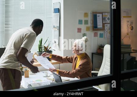 Multirassischer Mann und Seniorin, die Dokumente in zeitgenössischen Büros untersuchen, mit Diagrammen und Grafiken an den Wänden. Ideale Umgebung für geschäftliche Zusammenarbeit Stockfoto