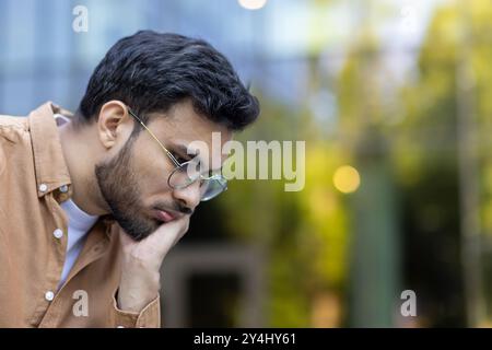 Nachdenklicher Mann mit Brille und braunem Hemd schaut nachdenklich nach unten und vermittelt Kontemplation und Konzentration. Die urbane Umgebung im Hintergrund verbessert die Stimmung der Introspektion und des Fokus. Stockfoto