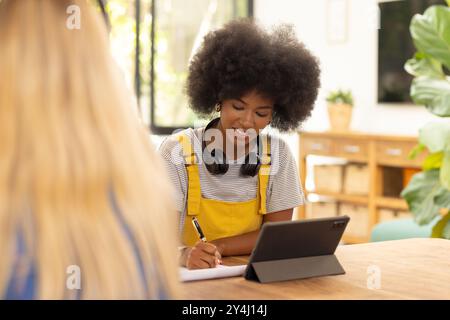 Schreiben in Notizblock, junge Frau, die ein Tablet mit Kopfhörern in ungezwungener Atmosphäre verwendet Stockfoto