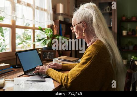 Ältere Frau mit langen weißen Haaren sitzt am Holzschreibtisch, benutzt Laptop und macht Notizen mit Papieren und Kaffeetasse herum, Pflanzen und Bücherregal im Hintergrund Stockfoto