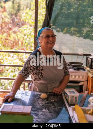 Alanya, Türkei - 17. September 2021: Eine ältere Frau auf einer Schürze lächelt, während sie in einer gemütlichen rustikalen Küche, umgeben von der Natur, Teig zubereitet Stockfoto