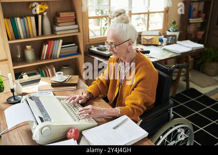 Eine ältere Frau, die im Rollstuhl sitzt und auf der Schreibmaschine in gut organisierter Büroumgebung mit Bücherregalen und Papieren um sie herum einen produktiven Arbeitsplatz zeigte Stockfoto