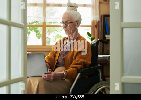 Ältere Frau, die im Rollstuhl sitzt und mit einem Laptop im Arbeitszimmer mit natürlichem Licht aus dem Fenster und Pflanzen im Hintergrund eine ruhige Atmosphäre schafft Stockfoto