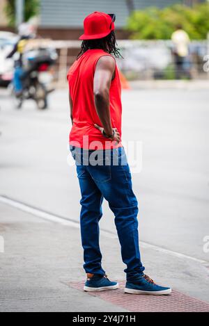 Schwarzer Mann mit rotem Hemd, rotem Hut und Jeans, der mit Hand auf Hüfte am Straßenrand in Belo Horizonte, Brasilien, steht. Stockfoto