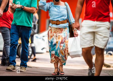 Brasilianische Frau mittleren Alters in blauer Bluse und farbenfrohem Rock mit Einkaufstasche, die in Savassi, Belo Horizonte, unter männlichen Fußgängern spazieren geht. Stockfoto