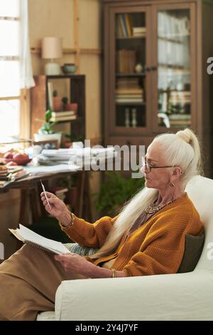 Seniorin sitzt im gemütlichen Arbeitszimmer, schreibt und denkt. Hintergrund voller Bücher, verschiedene Schreibtischgegenstände und eine warme Atmosphäre schaffen einen Stockfoto