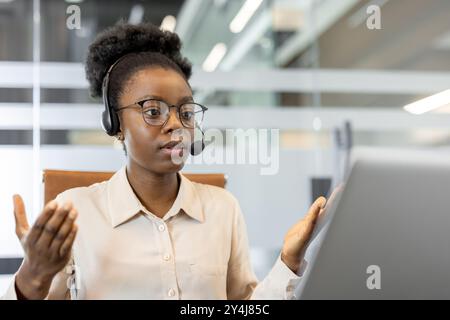 Junge afroamerikanische Frau im Büro, die ein Headset trägt, nimmt an professionellen Videoanrufen Teil. Sie sind an Diskussionen beteiligt und sprechen selbstbewusst mit Kollegen über ein Notebook Stockfoto