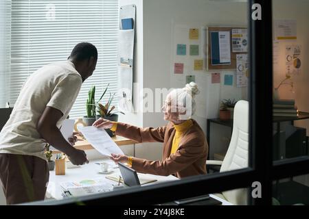 Multirassischer Mann und Seniorin, die Dokumente in zeitgenössischen Büros untersuchen, mit Diagrammen und Grafiken an den Wänden. Ideale Umgebung für geschäftliche Zusammenarbeit oder Teamarbeit Stockfoto