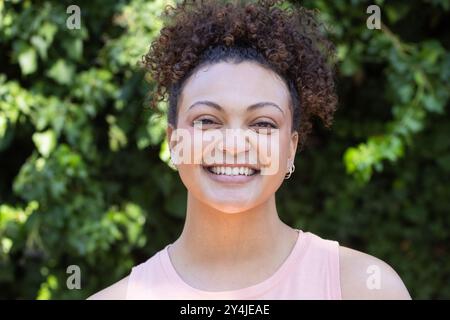 Lächelnde Frau mit lockigem Haar, die den Lebensstil im Freien genießt, vor grünem Laub stehend Stockfoto