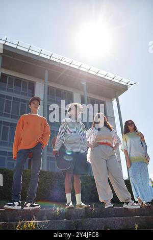 Vertikale Aufnahme von coolen Teenagern, die auf Stufen vor der Tür stehen und in einer Vierergruppe selbstbewusst in die Ferne schauen und legere Outfits zum Laufen zeigen Stockfoto