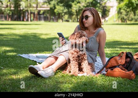 Frau, die Buch liest, entspannt sich auf Picknickteppich mit ihrem flauschigen braunen Maltipoo-Hund an sonnigen Tagen. Diese Outdoor-Szene fängt die Freude an der Bindung mit Haustieren und en ein Stockfoto