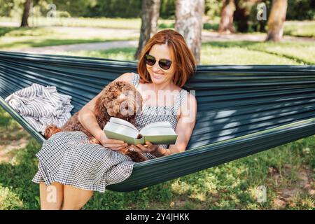 Frau, die Buch liest, entspannt sich in der Hängematte mit ihrem flauschigen braunen Maltipoo-Hund an sonnigem Tag. Sowohl zufrieden als auch glücklich. Diese Outdoor-Szene fängt Jo ein Stockfoto