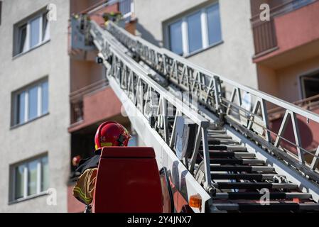 Der Bediener eines Feuerwehrwagens mit verlängerter Hochleiter reicht bis zum Wohngebäude, in dem Feuerwehrleute in Schutzausrüstung auf dem Balkon arbeiten Stockfoto