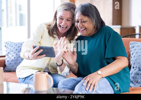 Zu Hause, lächelnde ältere, multirassische Freundinnen, Videoanrufe auf dem Smartphone, sitzen auf der Couch Stockfoto