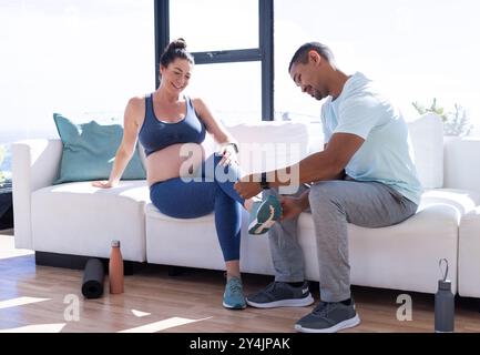 Zu Hause, multirassisches Paar, hilft schwangeren Frauen mit Schuhen, Mann lächelt und sitzt auf der Couch Stockfoto