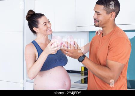 Zu Hause, schwangere, multirassische Paare, die mit Smoothies in der modernen Küche trösten, lächelnd Stockfoto