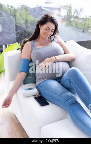 Zu Hause, schwangere Frau, die sich auf der Couch entspannt und den Blutdruck mit dem Gerät überwacht Stockfoto