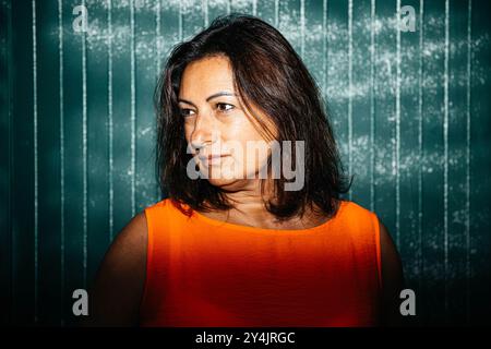 Das Frauenporträt trägt ein orangefarbenes Oberteil, Schatten auf grünem Hintergrund, zeigt nachdenklichen Ausdruck, emotionales Konzept Stockfoto