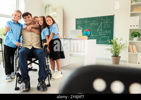 Männlicher Lehrer im Rollstuhl, der sich mit kleinen Schülern in der Schule umarmt Stockfoto