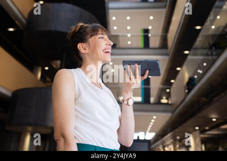 Lächelnde Geschäftsfrau mit Smartphone für Sprachnachrichten in modernen Bürogebäuden Stockfoto
