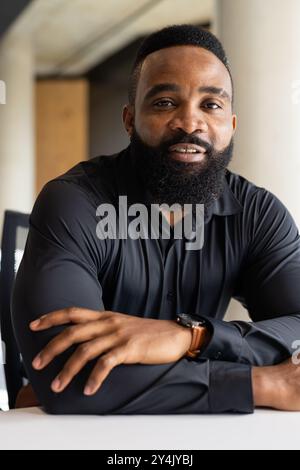 Lächelnder afroamerikanischer Geschäftsmann in schwarzem Hemd am Schreibtisch im modernen Büro Stockfoto