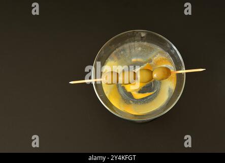 Ein Glas trockenen Wermut mit Zitronenschale und einem Spieß mit vier Oliven auf schwarzem Hintergrund. Draufsicht, flach. Stockfoto