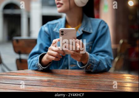 Ein zugeschnittenes Bild einer asiatischen Frau in einer Jeansjacke, die Kopfhörer trägt, an einem Restauranttisch sitzt, ihr Smartphone hält und aw aussieht Stockfoto