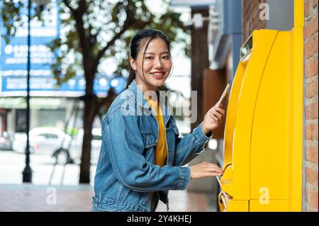 Eine schöne asiatische Frau in einer Jeansjacke steht vor einem Geldautomaten und lächelt in die Kamera. Geld abheben, Geldautomat, Personen und Stockfoto