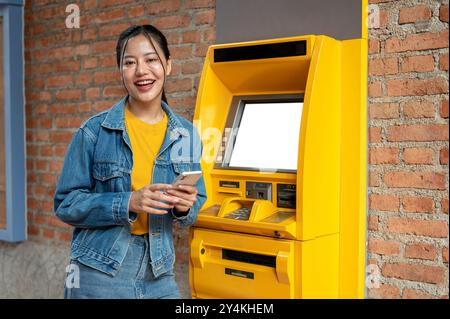 Eine schöne asiatische Frau in einer Jeansjacke steht vor einem Geldautomaten und lächelt in die Kamera. Geld abheben, Geldautomat, Personen und Stockfoto