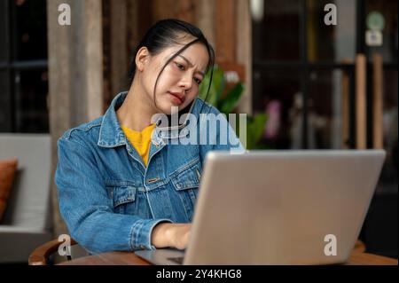 Eine gestresste, beschäftigte und unzufriedene asiatische Frau in einer Jeansjacke telefoniert mit ihrem Kollegen und beschwert sich über ein Problem, während sie an ihrem Laptop arbeitet Stockfoto