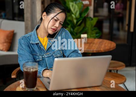 Eine gestresste, beschäftigte und unzufriedene asiatische Frau in einer Jeansjacke telefoniert mit ihrem Kollegen und beschwert sich über ein Problem, während sie an ihrem Laptop arbeitet Stockfoto