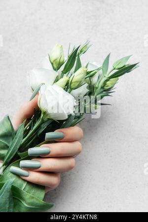 Eine zarte Hand, die mit langen grünen Nägeln verziert ist, umhüllt sanft einen Blumenstrauß aus weißen Blumen und zeigt einen ruhigen Kontrast vor einem weichen grauen Hintergrund. Stockfoto