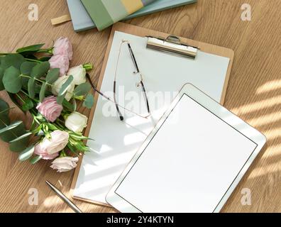 Ein heller Arbeitsbereich mit leerem Tablet, offenem Notizbuch, Schreibutensilien und frischen Blumen auf einem Holztisch lädt zu Kreativität und Kreativität ein Stockfoto