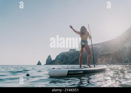 Frau Paddleboarding Ocean Coastline - Frau genießt an einem sonnigen Tag eine Paddleboard-Session auf dem Meer mit felsigen Klippen im Hintergrund. Stockfoto