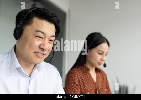 Asiatische Büroangestellte mit Headsets konzentrierten sich auf Telekommunikationsaufgaben. Männliche und weibliche Fachleute arbeiten in modernen Büroumgebungen zusammen und nutzen das Telefon für Kundenservice oder technischen Support. Stockfoto