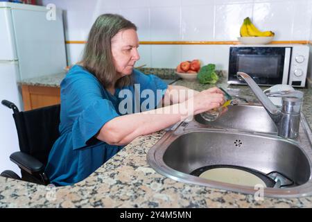 Reife Frau im Rollstuhl, die Geschirr in der Küche zu Hause wäscht Stockfoto