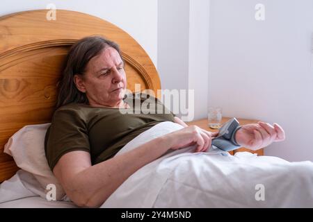 Frau über 60 Jahre, die ihren Blutdruck im Bett zu Hause mit einem Handgelenktendiometer misst Stockfoto