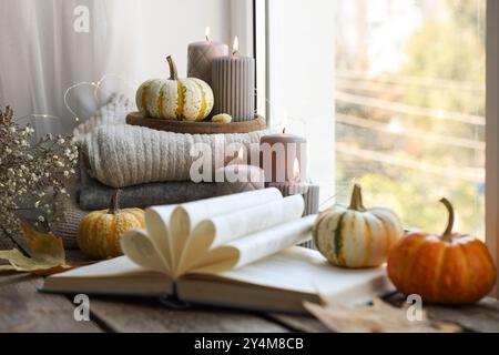 Offenes Buch, Stapel Pullover, brennende Kerzen und Kürbisse auf der Fensterbank. Herbstatmosphäre Stockfoto