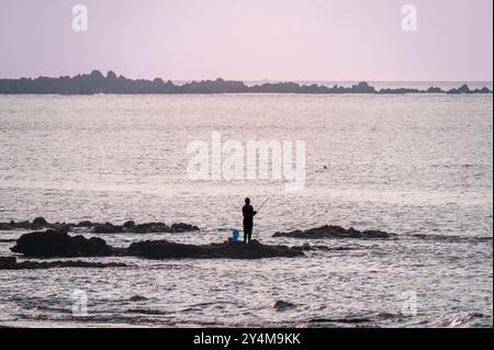 Ein einsamer Fischer, der auf einem Felsvorsprung vor einem atemberaubenden Sonnenuntergang steht. Die Silhouette des Fischers schafft ein Gefühl der Ruhe und Einsamkeit. S Stockfoto