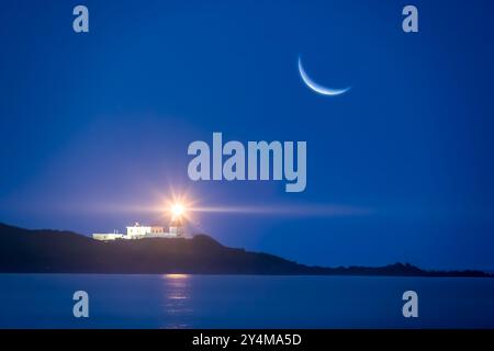 Ein Halbmond erleuchtet eine ruhige nächtliche Meereslandschaft und strahlt sein sanftes Leuchten über einen Leuchtturmstrahl. Die ruhige Szene ist perfekt zum Entspannen und Stockfoto