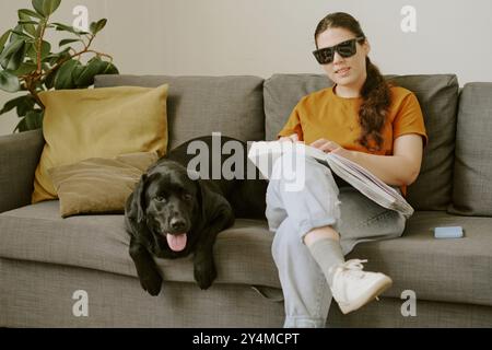 Junge Frau mit Behinderung, die auf dem Sofa sitzt und Blindenschrift liest, während ihr Haustier neben ihr liegt Stockfoto