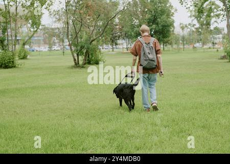 Rückansicht eines blinden Mannes, der mit schwarzem Blindenhund im grünen Park aufwacht Stockfoto