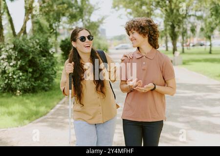 Zwei beste Freunde, die laut lachen, während sie im grünen Park spazieren, eine brünette junge Frau, die Stock benutzt, um sich zu orientieren Stockfoto