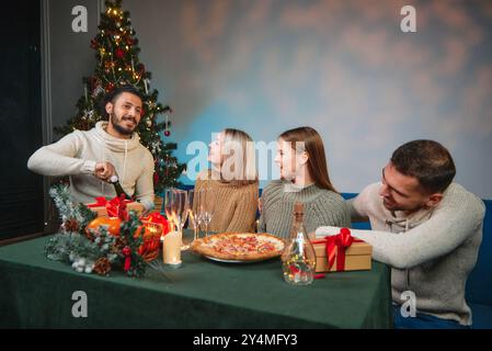 Vier Freunde sitzen an einem Tisch mit Pizza und Getränken, eröffnen Geschenke und feiern gemeinsam Weihnachten. Stockfoto