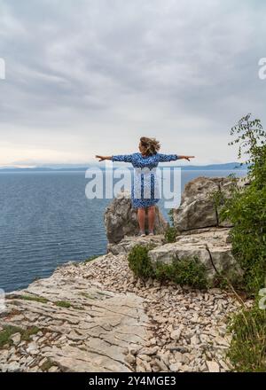 Glückliche Frau mittleren Alters genießt mit ausgestreckten Armen auf dem Abgrund der felsigen Küste und blickt auf die Meere Stockfoto